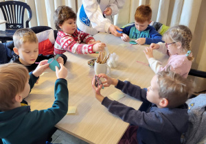 Sześcioro dzieci siedzi przy stoliku, dekorują drewniane bombki, pomaga im pani przebrana za śnieżynkę