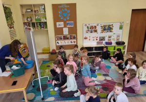 Dzieci siedzą na dywanie, odgadują i nasłuchują dźwięki wytwarzane przez nauczyciela.