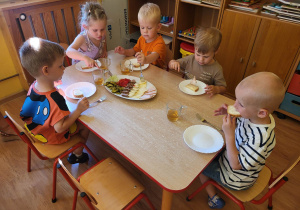 Dzieci siedzą przy stole i częstują się produktami spożywczymi leżącymi na wspólnej tacy.