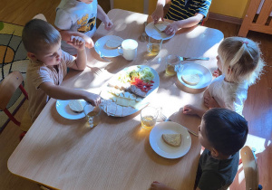 Dzieci siedzą przy stole i częstują się produktami spożywczymi leżącymi na wspólnej tacy.