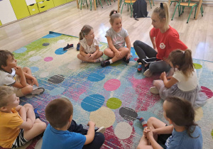 Dzieci siedzą na dywanie z instruktorem.