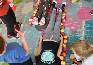 Na dywanie leży dwoje dzieci, wzdłuż nich ułożone są jabłka