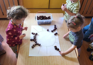 Dzieci układają z kasztanow wzory. Na zdjęciu: Pola, Tymek i Marianna układają kasztany na konturze słonka.
