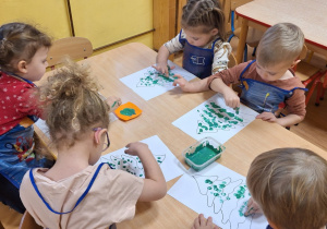 Dzieci stemplują palcami umoczonym w zielonej farbie plakatowej na kartce z konturem choinki.