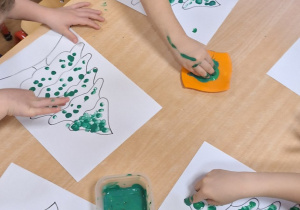 Dzieci stemplują palcami umoczonym w zielonej farbie plakatowej na kartce z konturem choinki.