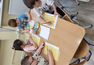 Dzieci siedzą przy stoliku, malują farbami na płatach lasagne