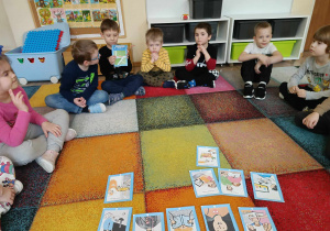 Dzieci siedzą na dywanie w kręgu, omawiają prezentowane ilustracje.