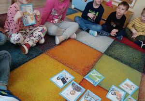 Dzieci siedzą na dywanie w kręgu, omawiają prezentowane ilustracje.