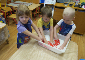 Hania, Antek i Filip mieszają masę z czerwonym barwnikiem.