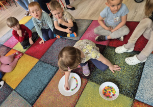 Dzieci siedzą na dywanie w kole, dziewczynka układa na talerzu cukierki