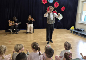 Dzieci siedzą w sali, przed nimi stoi muzyk, w tle dwoje muzyków gra na gitarze i altówce