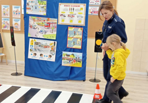 Dziewczynka przechodzi po makiecie przejścia dla pieszych, w asyście policjantki.