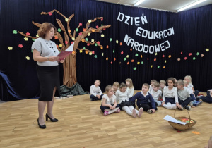 Dzieci siedzą na scenie, obok nich stoi pani dyrektor.