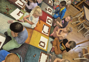 Dzieci siedzą na dywanie, układają kasztany na kartkach papieru