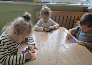 Dzieci siedzą przy stoliku, wydrapują wzorki na kartoniku pokrytym plasteliną