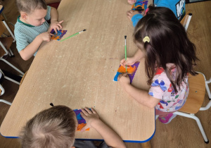 Dzieci siedzą przy stoliku, wydrapują wzorki na kartoniku pokrytym plasteliną