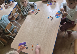Dzieci siedzą przy stoliku, rozcierają plastelinę na kartoniku