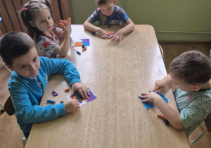 Dzieci siedzą przy stoliku, rozcierają plastelinę na kartoniku