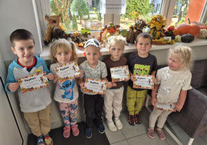 Uśmiechnięte dzieci z dyplomami w ręku stoją przed pracami konkursowymi