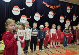 Dzieci stoją na scenie, jedna dziewczynka mówi do mikrofonu