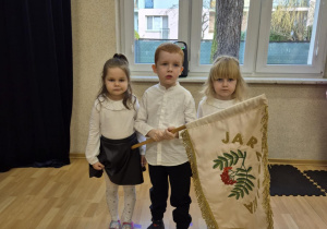 Chłopiec trzyma proporzec przedszkola, przy nim stoją dwie dziewczynki