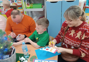 Rodzice z dziećmi siedzą przy stolikach, ozdabiają bombki.