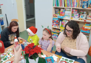 Rodzice z dziećmi siedzą przy stolikach, ozdabiają bombki.