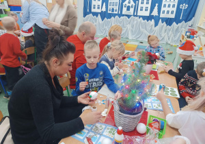 Rodzice z dziećmi siedzą przy stolikach, ozdabiają bombki.