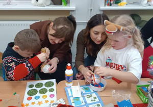 Rodzice z dziećmi siedzą przy stolikach, ozdabiają bombki.