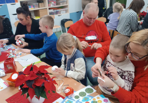 Rodzice z dziećmi siedzą przy stolikach, ozdabiają bombki oraz kartki świąteczne.