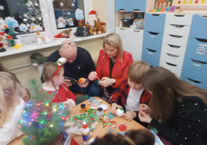 Rodzice z dziećmi siedzą przy stolikach, ozdabiają bombki.