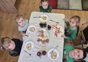 Dzieci siedzą przy stole nakrytym obrusem i pozują do zdjęcia. Na stole stoją talerze z jedzeniem.
