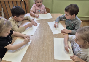 Dzieci siedzą przy stolikach, malują świecami na kartkach papieru