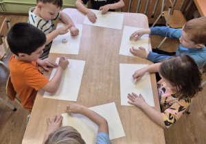 Dzieci siedzą przy stolikach, malują świecami na kartkach papieru