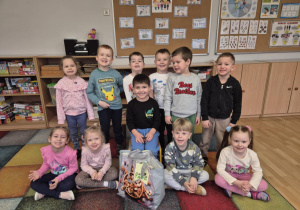 Grupa dzieci siedzi na dywanie wokół chłopca, który siedzi na krzesełku.