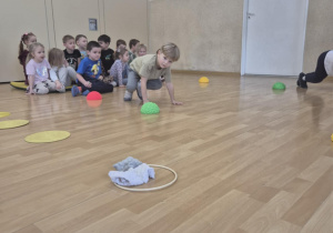 Chłopiec ćwiczy w pozycji na czworaka, za chłopcem siedzą dzieci z rzędzie.