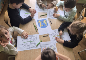 Sześcioro dzieci siedzi przy stoliku i koloruje herb Kutna.