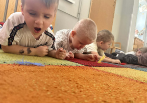 Dzieci leżą na dywanie i dmuchają na piórka.