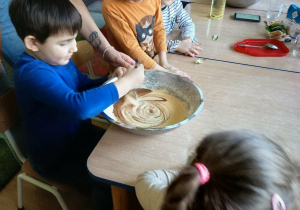 Dzieci siedzą przy stolikach, chłopiec miesza łyżką ciasto