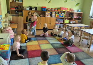 Dzieci siedzą w kole na dywanie, przed nimi kobieta czyta książkę.