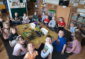 Dzieci siedzą w kole. Na środku znajdują się przyniesione przez rodziców i dzieci kwiaty, cebulki, nasiona, doniczki, konewka i ziemia.
