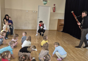 Jeden z muzyków gra na fagocie, drugi razem z dziećmi kuca na podłodze
