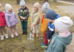 Dzieci stoją na trawniku, jedna z dziewczynek trzyma grabie