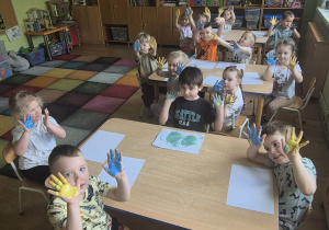 Dzieci siedzą przy stolikach, pokazują dłonie pomalowane na żółto i niebiesko