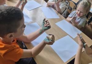 Uśmiechnięte dzieci siedzą przy stolikach, rozcierają farby pomiędzy dłońmi