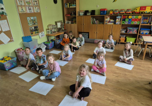 Dzieci siedzą na podłodze, przed nimi leżą arkusze białego papieru