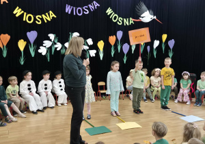 Dzieci w różnych przebraniach na tle wiosennej dekoracji, przed nimi stoi nauczycielka oraz chłopiec z tabliczką