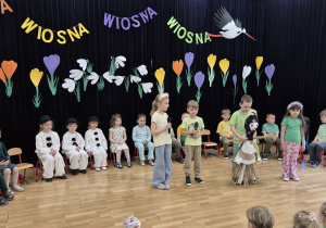 Dzieci w różnych przebraniach na tle wiosennej dekoracji, jeden z chłopców trzyma Marzannę