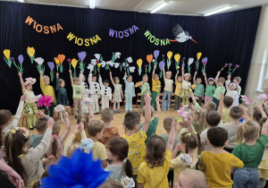 Dzieci w różnych przebraniach na tle wiosennej dekoracji trzymają w rękach kwiaty, przed nimi dzieci na widowni machają kwiatami