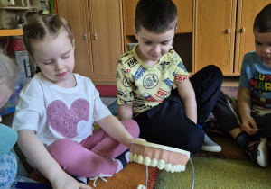 Dzieci siedzą na dywanie, dziewczynka na modelu zębów prezentuje właściwy sposób ich mycia